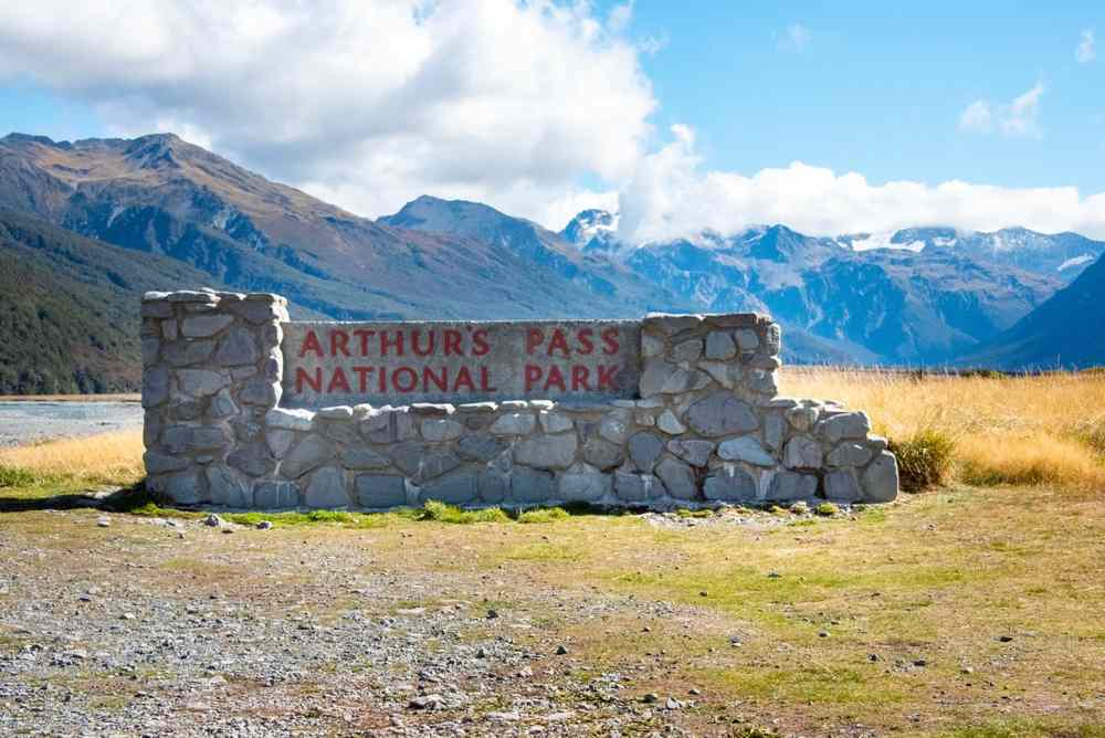 Stone entrance sign for Arthur’s Pass National Park with the Southern Alps rising behind it on New Zealand’s South Island, bright day and wide valley views.