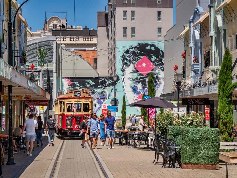 Christchurch city street with the iconic red tram, pedestrians, cafés, and a large mural, capturing the lively downtown vibe on a sunny day.