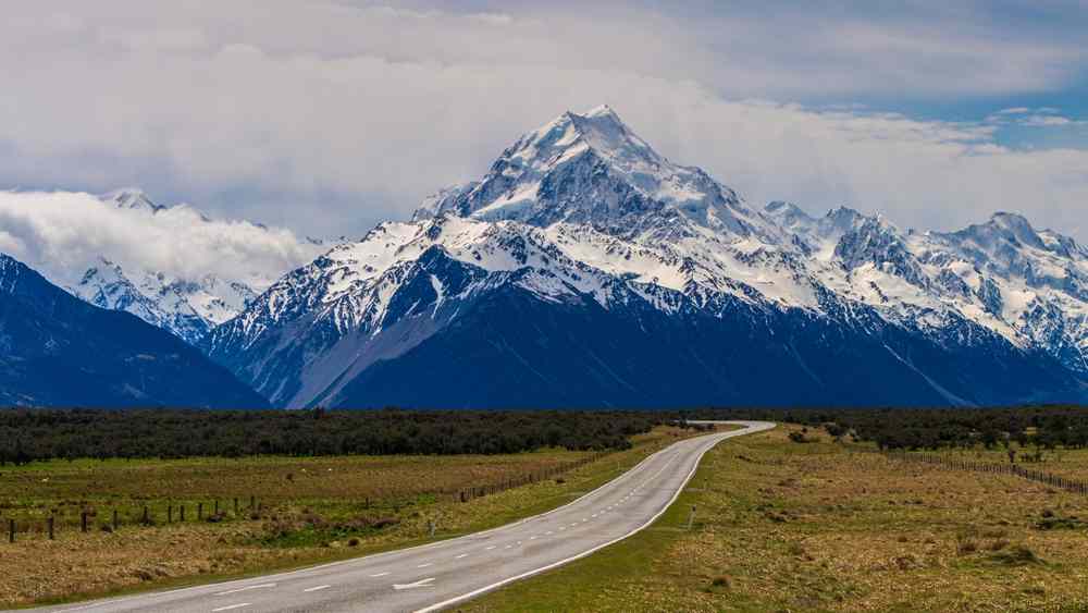 Open road leading toward Aoraki/Mount Cook, with rugged snowcapped peaks ahead and wide South Island plains on either side under moody clouds.
