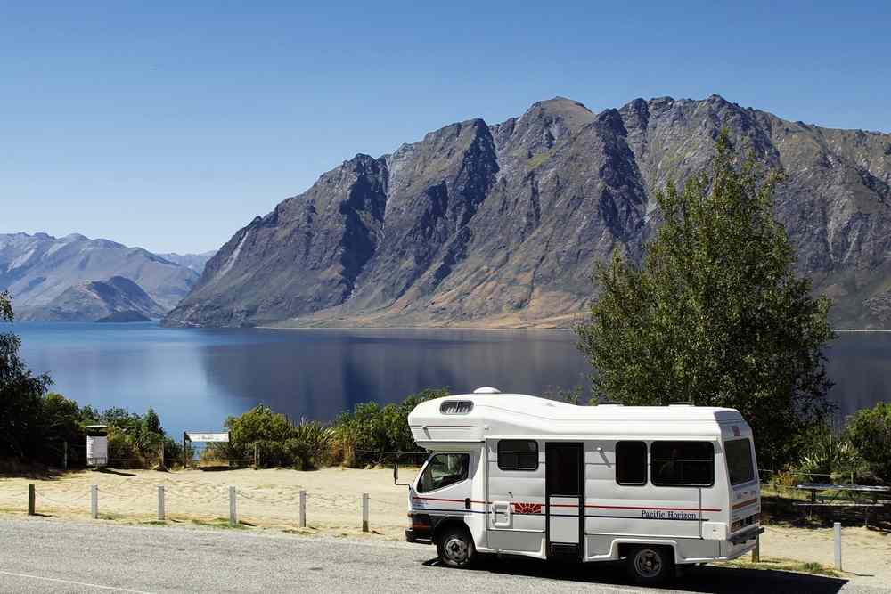 Motorhome parked by Lake Wānaka with towering mountains across the water, classic South Island camper trip scene on a bright, calm day.